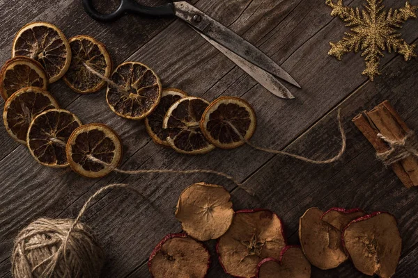 Vue de dessus des tranches d'agrumes et de pommes séchées près du flocon de neige, de la cannelle, de la corde et des ciseaux sur la surface en bois — Photo de stock