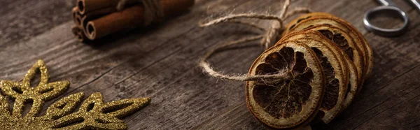 Selective focus of dried citrus slices on thread near cinnamon, scissors and snowflake on wooden background, panoramic shot — Stock Photo