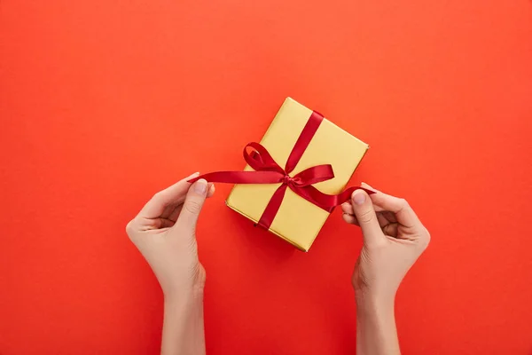 Cropped view of woman untying ribbon on golden gift box on red background — Stock Photo