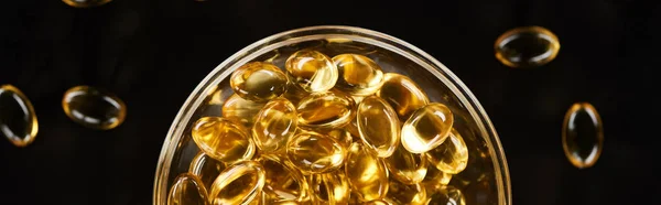 Vue du dessus des capsules d'huile de poisson dorées dans un bol en verre isolé sur plan panoramique noir — Photo de stock