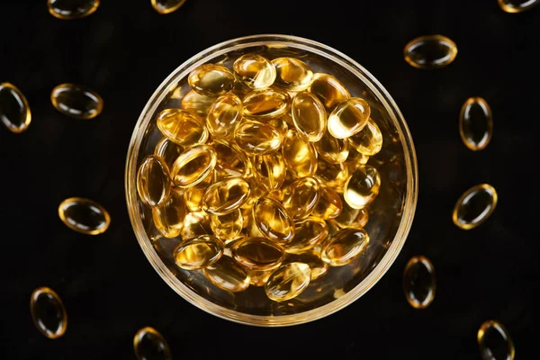 Vue du dessus des capsules d'huile de poisson dorées dans un bol en verre isolé sur noir — Photo de stock