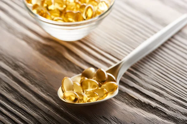 Mise au point sélective de capsules d'huile de poisson dorées dans une cuillère et un bol en verre sur une table en bois — Photo de stock