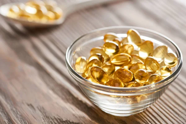 Foyer sélectif de capsules d'huile de poisson doré dans un bol en verre et cuillère sur table en bois — Photo de stock