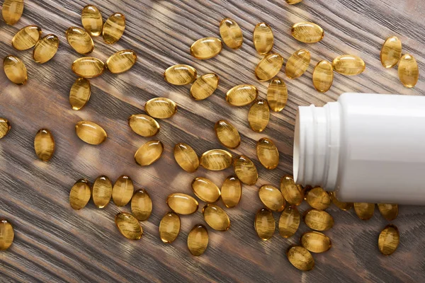 Capsule di olio di pesce dorato sparse dal contenitore di plastica sul tavolo di legno — Foto stock