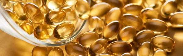 Close up view of shiny fish oil capsules scattered from glass bowl on golden background, panoramic shot — Stock Photo