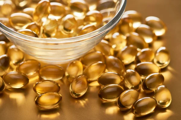 Close up view of shiny fish oil capsules scattered from glass bowl on golden background — Stock Photo