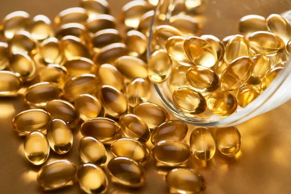 Close up view of shiny fish oil capsules scattered from glass bowl on golden background — Stock Photo