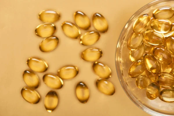 Top view of shiny golden fish oil capsules scattered from glass bowl on yellow background — Stock Photo