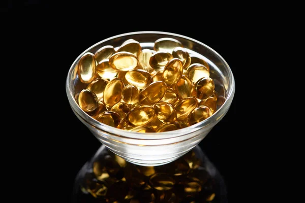 Golden shiny fish oil capsules in glass bowl on black background — Stock Photo