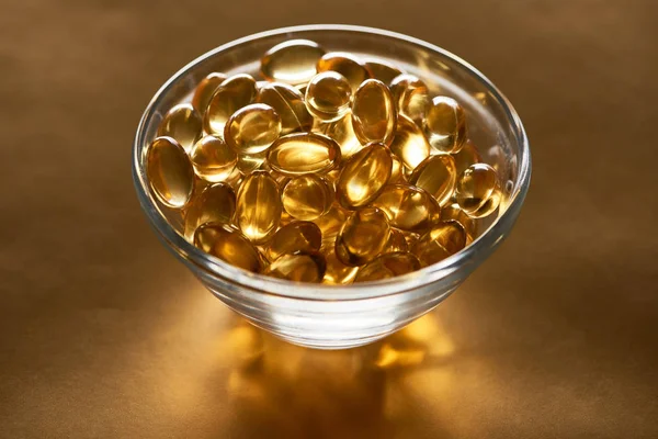 Cápsulas de óleo de peixe brilhante em tigela de vidro sobre fundo dourado — Fotografia de Stock