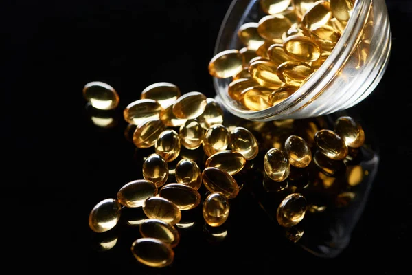 Shiny golden fish oil capsules scattered from glass bowl on black background — Stock Photo