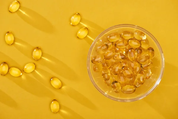 Vista superior de cápsulas de aceite de pescado brillante dorado dispersas cerca de un recipiente de vidrio sobre un fondo amarillo brillante - foto de stock