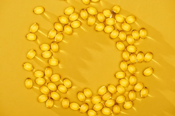 Vue de dessus des capsules d'huile de poisson brillantes dorées disposées en cercle sur fond lumineux jaune avec espace de copie — Photo de stock