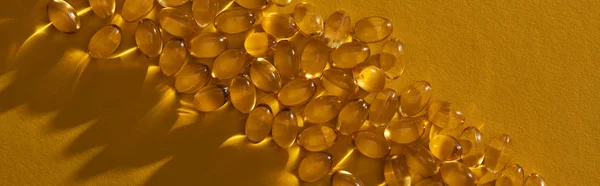 Vista superior de cápsulas de aceite de pescado brillante dorado sobre fondo amarillo en plano oscuro y panorámico - foto de stock