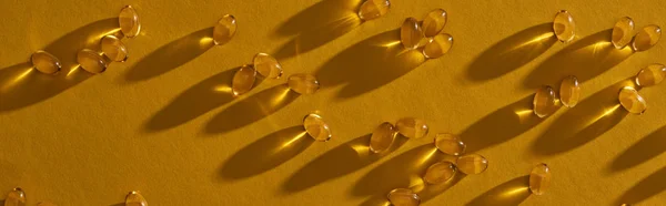 Top view of golden shiny fish oil capsules scattered on yellow background in dark, panoramic shot — Stock Photo