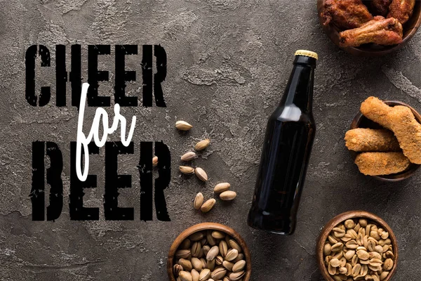 Top view of bottle of dark beer near bowls with snacks on grey surface with cheer for beer lettering — Stock Photo