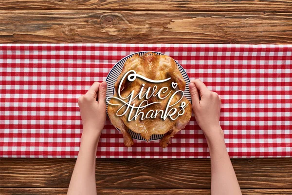 Cropped view of woman holding roasted turkey with give thanks illustration on red plaid napkin on wooden table — Stock Photo