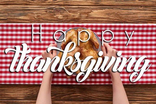 Cropped view of woman holding roasted turkey on red plaid napkin on wooden table with happy thanksgiving illustration — Stock Photo
