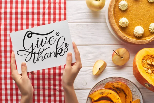 Partial view of woman holding card with give thanks illustration near pumpkin pie on wooden white table with apples and plaid red napkin — Stock Photo