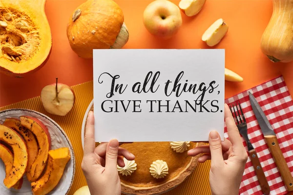 Cropped view of woman holding card with in all things give thanks illustration near pumpkin pie on orange background — Stock Photo