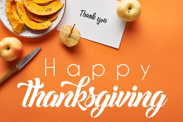 Vista superior de calabaza, manzanas, cuchillo y tarjeta de agradecimiento sobre fondo naranja con feliz ilustración de acción de gracias - foto de stock