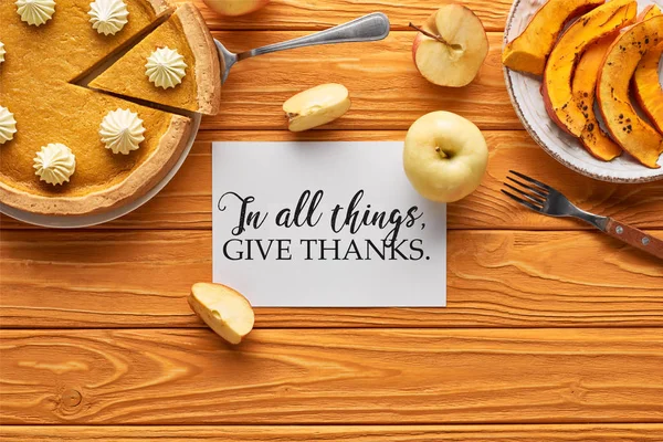 Vista superior de delicioso pastel de calabaza, manzanas y tarjeta con en todas las cosas dar gracias ilustración en mesa naranja de madera - foto de stock