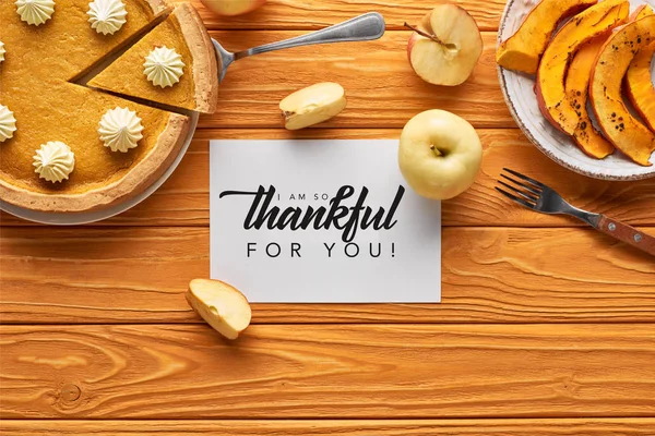 Top view of delicious pumpkin pie, apples and card with i am so thankful for you illustration on wooden orange table — Stock Photo