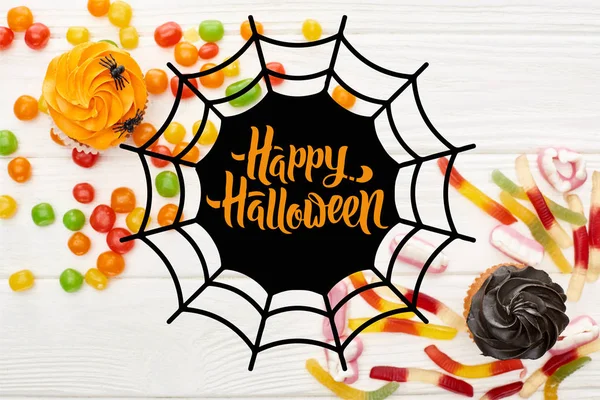 Top view of colorful gummy sweets, cupcakes and bonbons on white wooden table with spiderweb and happy Halloween illustration — Stock Photo