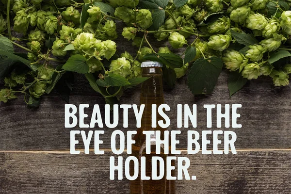 Top view of fresh beer in bottle with green hop on wooden surface with beauty is in the eye of the beer holder illustration — Stock Photo