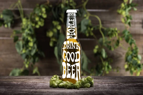 Fresh beer in bottle with good people drink good beer lettering near green hop on wooden surface — Stock Photo