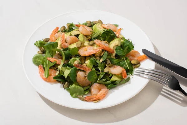 Frischer grüner Salat mit Kürbiskernen, Garnelen und Avocado auf Teller neben Besteck auf weißem Hintergrund — Stockfoto