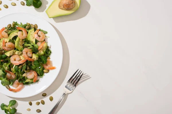 Vista dall'alto di insalata verde fresca con semi di zucca, gamberetti e avocado sul piatto vicino alla forchetta e ingredienti su sfondo bianco — Foto stock