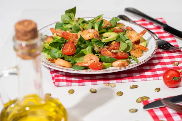 Selective focus of fresh green salad with shrimps and avocado on plate near cutlery on plaid napkin and jar of oil on white background — Stock Photo