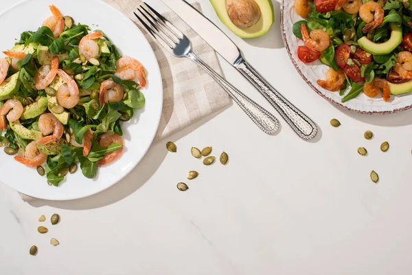 Draufsicht auf frischen grünen Salat mit Garnelen und Avocado auf Tellern in der Nähe von Besteck auf weißem Hintergrund — Stockfoto