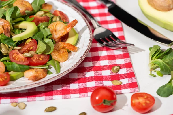 Vista ravvicinata di insalata verde fresca con gamberetti e avocado su piatto vicino a posate su tovagliolo plaid e ingredienti su sfondo bianco — Foto stock