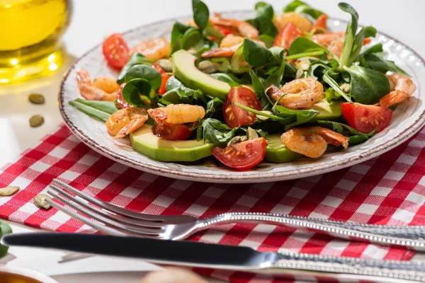 Enfoque selectivo de ensalada verde fresca con camarones y aguacate en el plato cerca de cubiertos en servilleta a cuadros - foto de stock