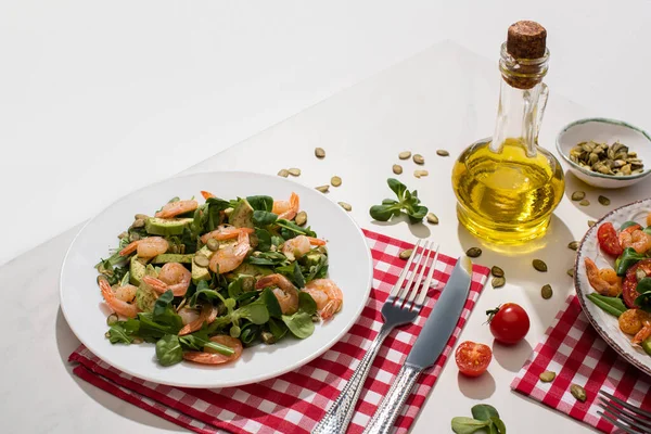 Свежий зеленый салат с креветками и авокадо на тарелке возле столовых приборов на клетчатой салфетке и ингредиенты на белом столе — стоковое фото
