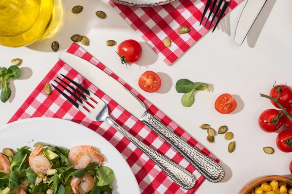 Vista dall'alto di insalata verde fresca con gamberetti e avocado su piatto vicino a posate su tovagliolo a quadri e ingredienti su sfondo bianco — Foto stock