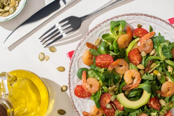 Top view of fresh green salad with shrimps and avocado on plate near cutlery on plaid napkin and ingredients on white background — Stock Photo