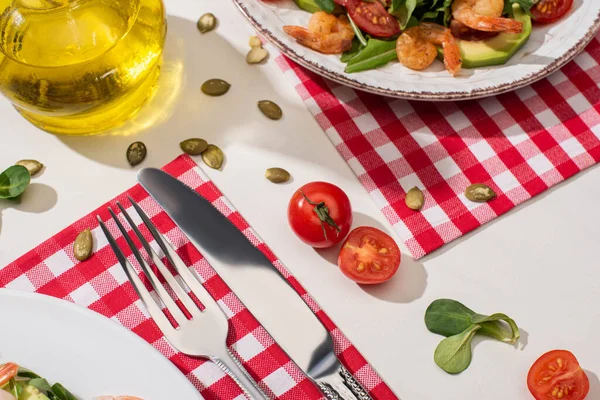 Свежий зеленый салат с креветками и авокадо на тарелке возле столовых приборов на клетчатой салфетке и ингредиенты на белом фоне — стоковое фото