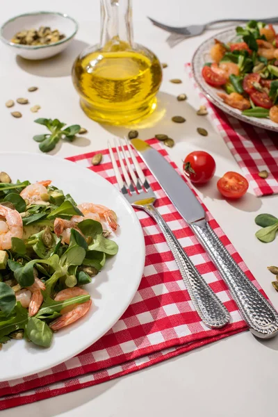 Fresh green salad with shrimps and avocado on plate near cutlery on plaid napkin and ingredients on white background — Stock Photo