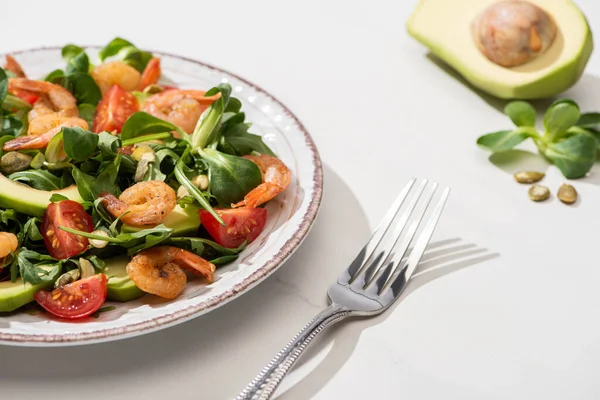 Foco seletivo de salada verde fresca com camarões e abacate na placa perto de garfo e ingredientes em fundo branco — Fotografia de Stock