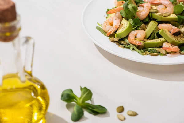 Selektiver Schwerpunkt von frischem grünem Salat mit Garnelen und Avocado auf Teller in der Nähe von Olivenöl auf weißem Hintergrund — Stockfoto