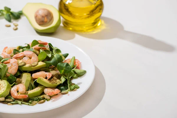 Selektiver Schwerpunkt von frischem grünem Salat mit Garnelen und Avocado auf Teller in der Nähe von Olivenöl auf weißem Hintergrund — Stockfoto