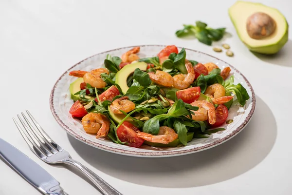 Foco seletivo de salada verde fresca com camarões e abacate na placa perto de talheres em fundo branco — Fotografia de Stock