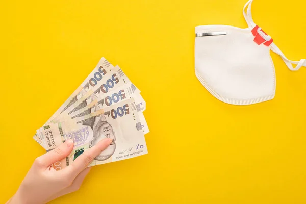 Partial view of woman holding Ukrainian banknotes near safety mask on yellow background — Stock Photo
