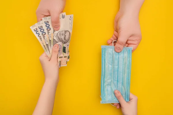 KYIV, UKRAINE - MARCH 25, 2020: partial view of two people holding Ukrainian banknotes and medical mask on yellow background — Stock Photo