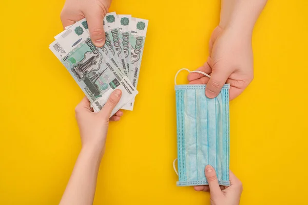 KYIV, UKRAINE - MARCH 25, 2020: partial view of two people holding Ukrainian banknotes and medical mask on yellow background — Stock Photo