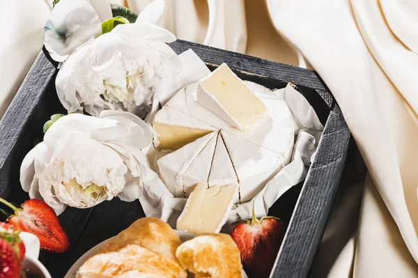 Nahaufnahme des französischen Frühstücks mit Croissant, Camembert, Erdbeeren auf Holztablett auf strukturiertem weißen Tuch mit Pfingstrosen — Stockfoto