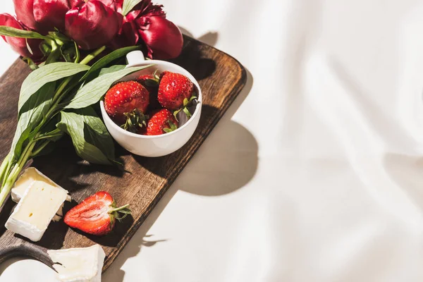 French breakfast with Camembert, strawberries and peonies on wooden cutting board on white tablecloth — Stock Photo